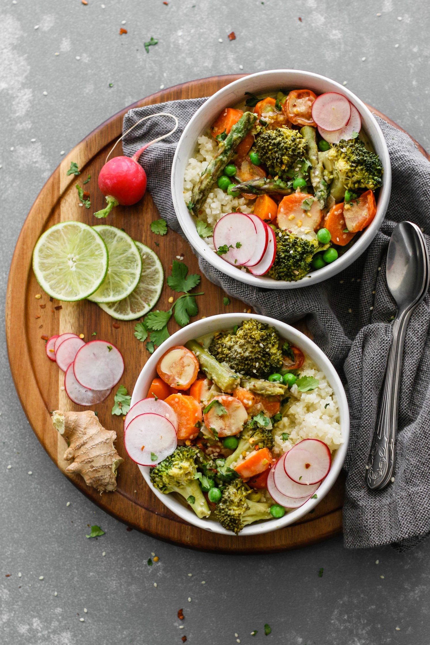 Green Coconut Curry with Cauliflower Rice served on wooden circle board with fresh lime, sliced radish, cilantro, and two spoons on the side