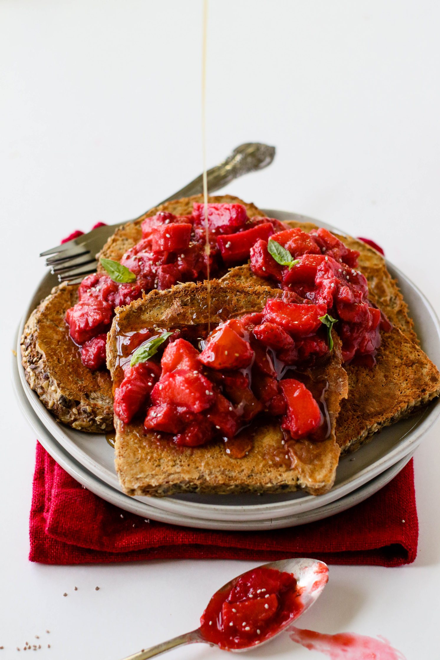 Strawberry Rhubarb French Toast served on white plate on red towel topped with strawberry rhubarb compote and fresh mint with two forks with maple syrup poured on top