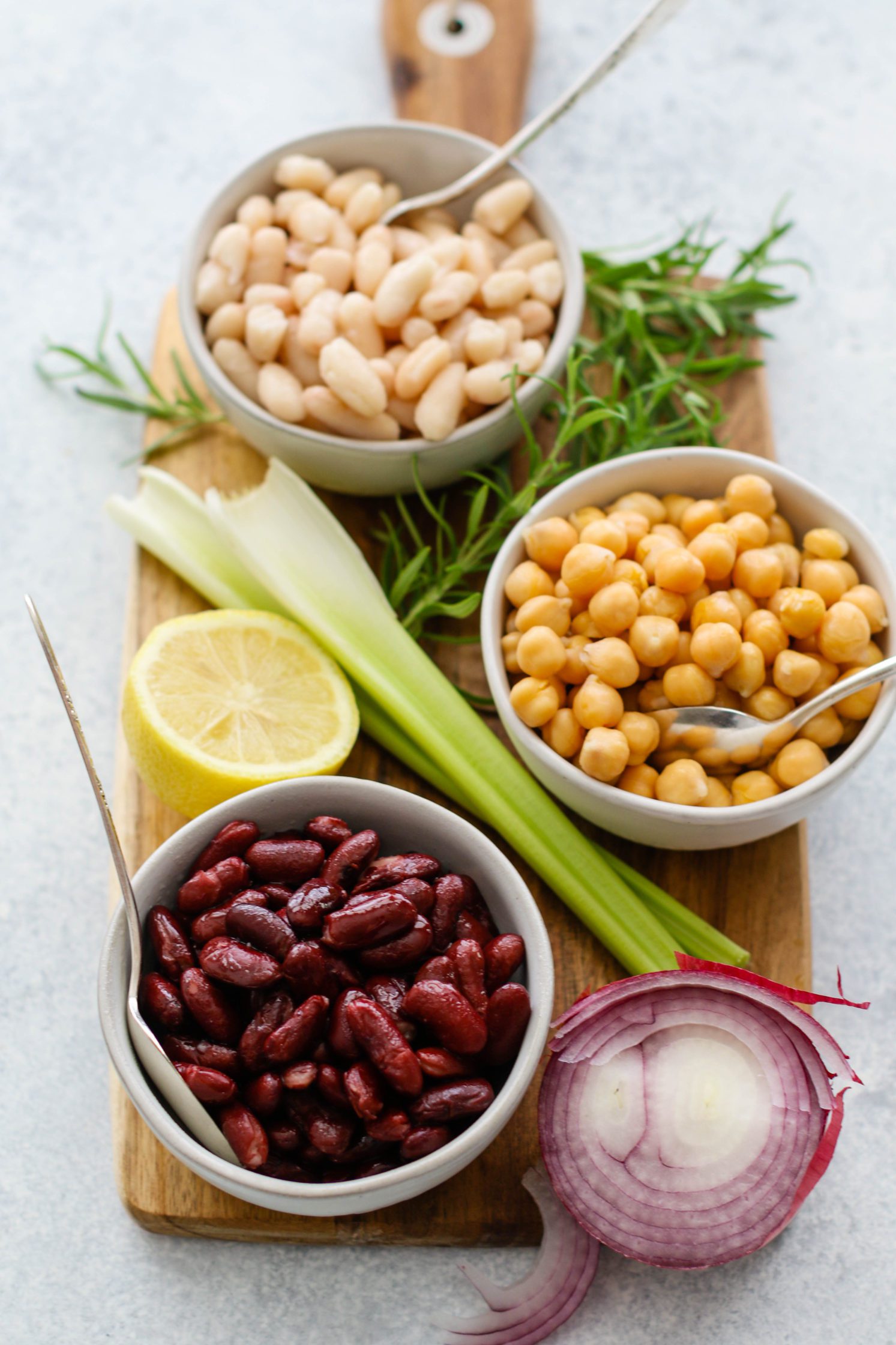 cannelini beans, chickpeas, and kidney beans in bowls with celery, lemon, red onion, and rosemary on wooden board