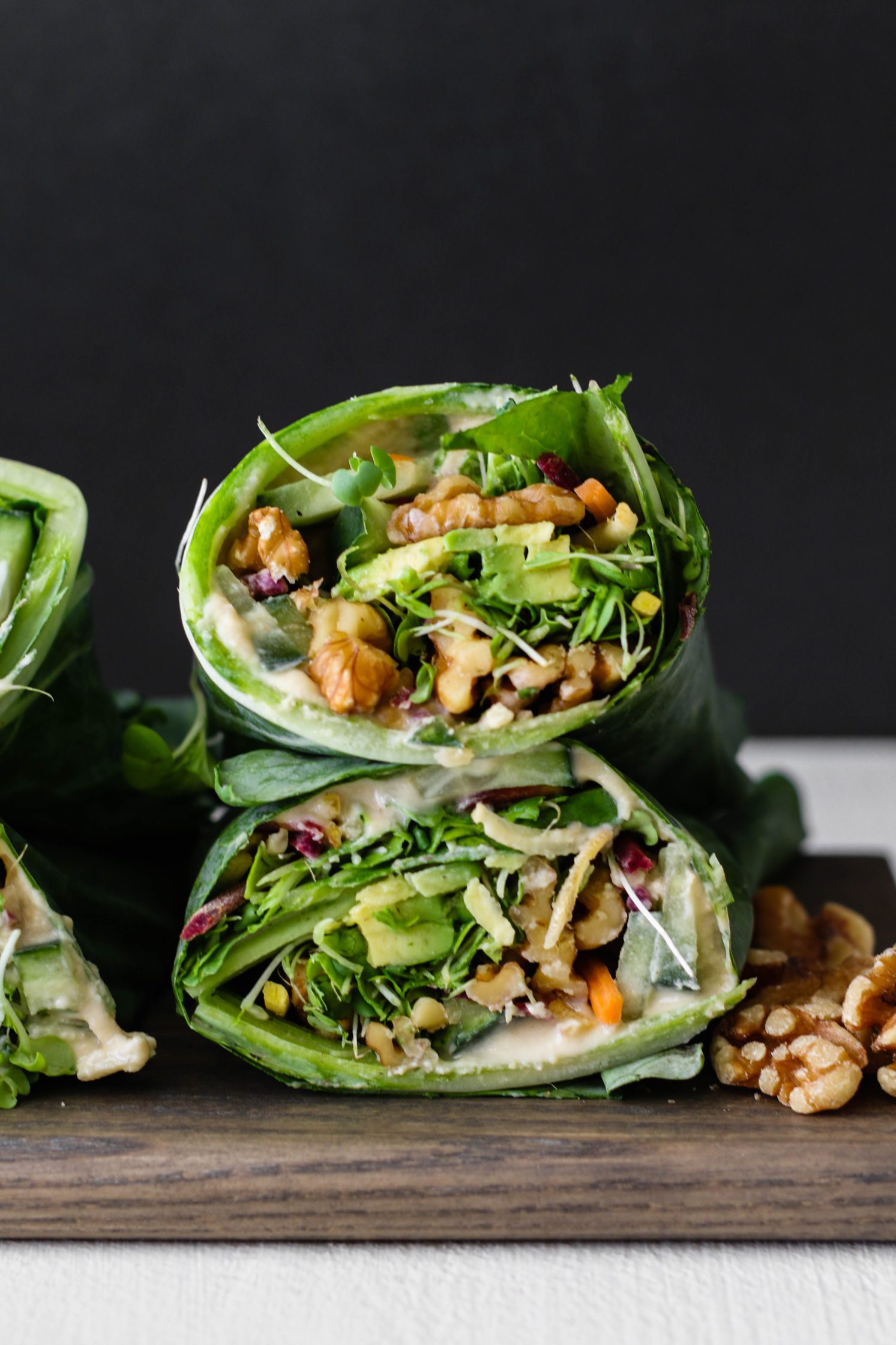 Rainbow Hummus & Avocado Collard Wraps stacked on wooden board with crushed walnuts by Flora & vino
