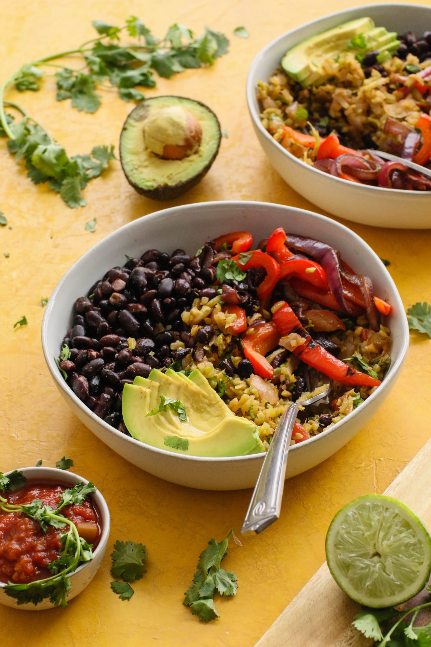  Broccoli Lime Rice & Black Bean Burrito Bowls served in bowls with forks topped with cilantro and lime and salsa and avocado on yellow board by Flora & Vino 