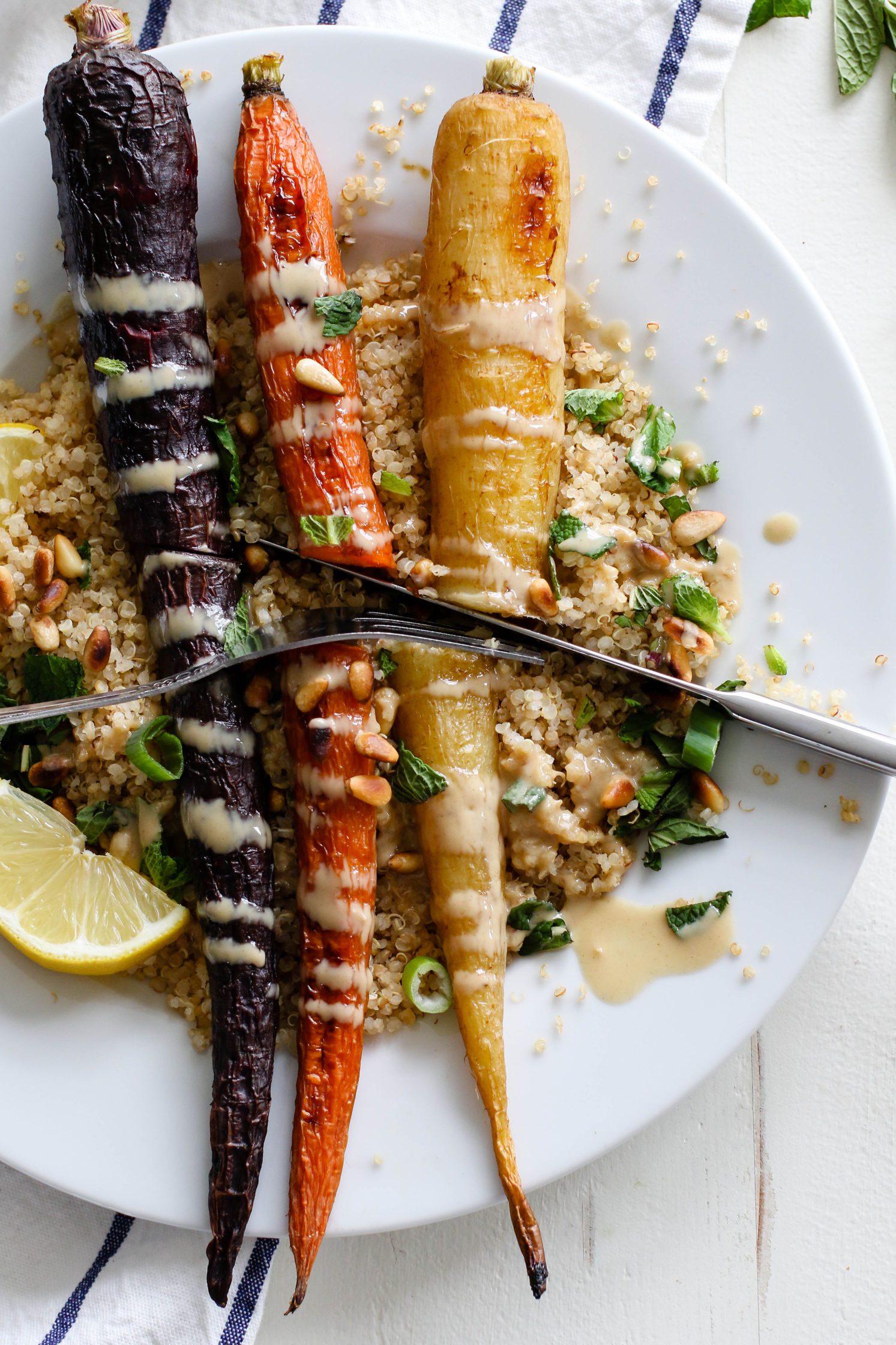 Rainbow Roasted Carrots served on plates over quinoa with fresh lemon, shredded mint, toasted pine nuts, and drizzle of tahini with fork and knife cutting into carrots