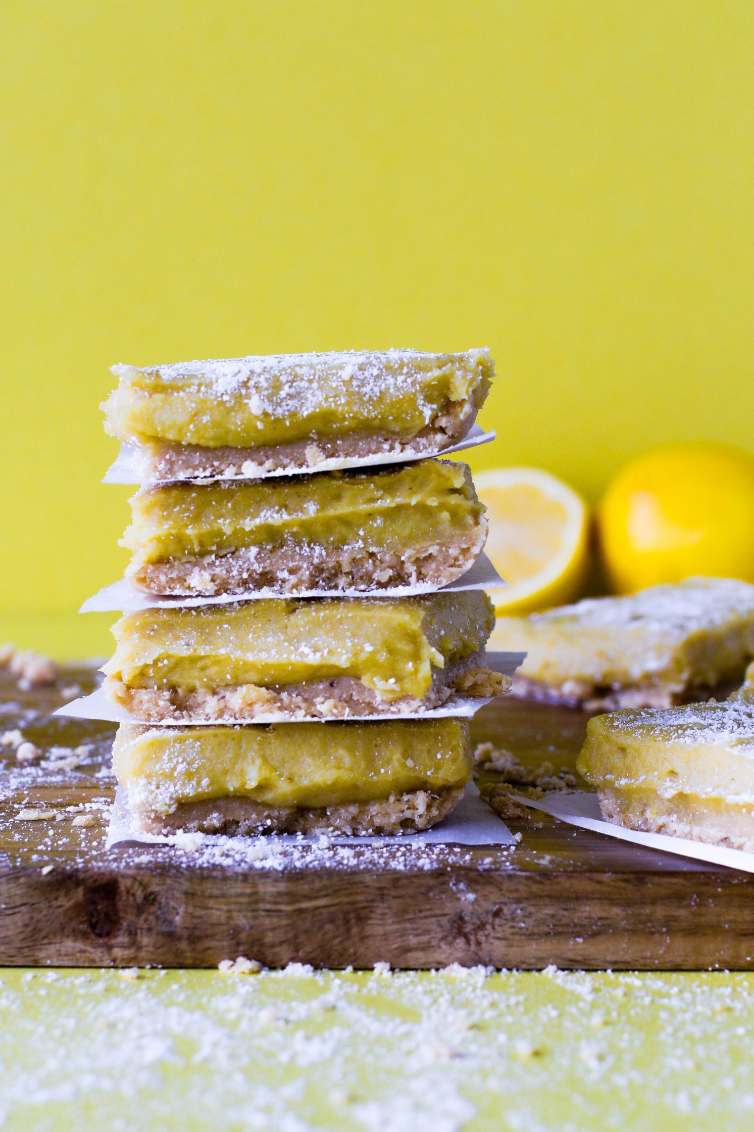 No-Bake Lemon Pie Bars stacked on wooden board with yellow background topped with coconut flour with lemons