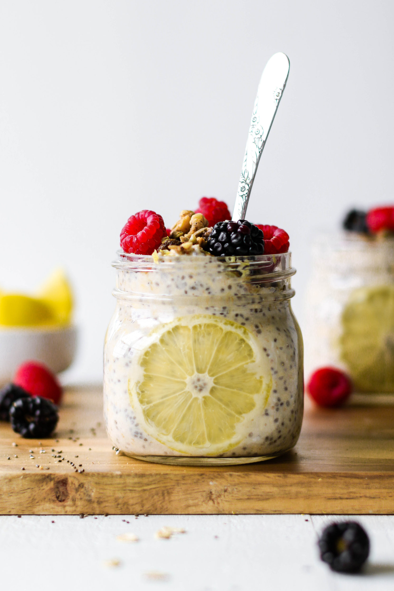 Lemon Berry Overnight Oats served in mason jars on a wooden board topped with fresh berries and lemon wedges with a spoon by Flora & Vino