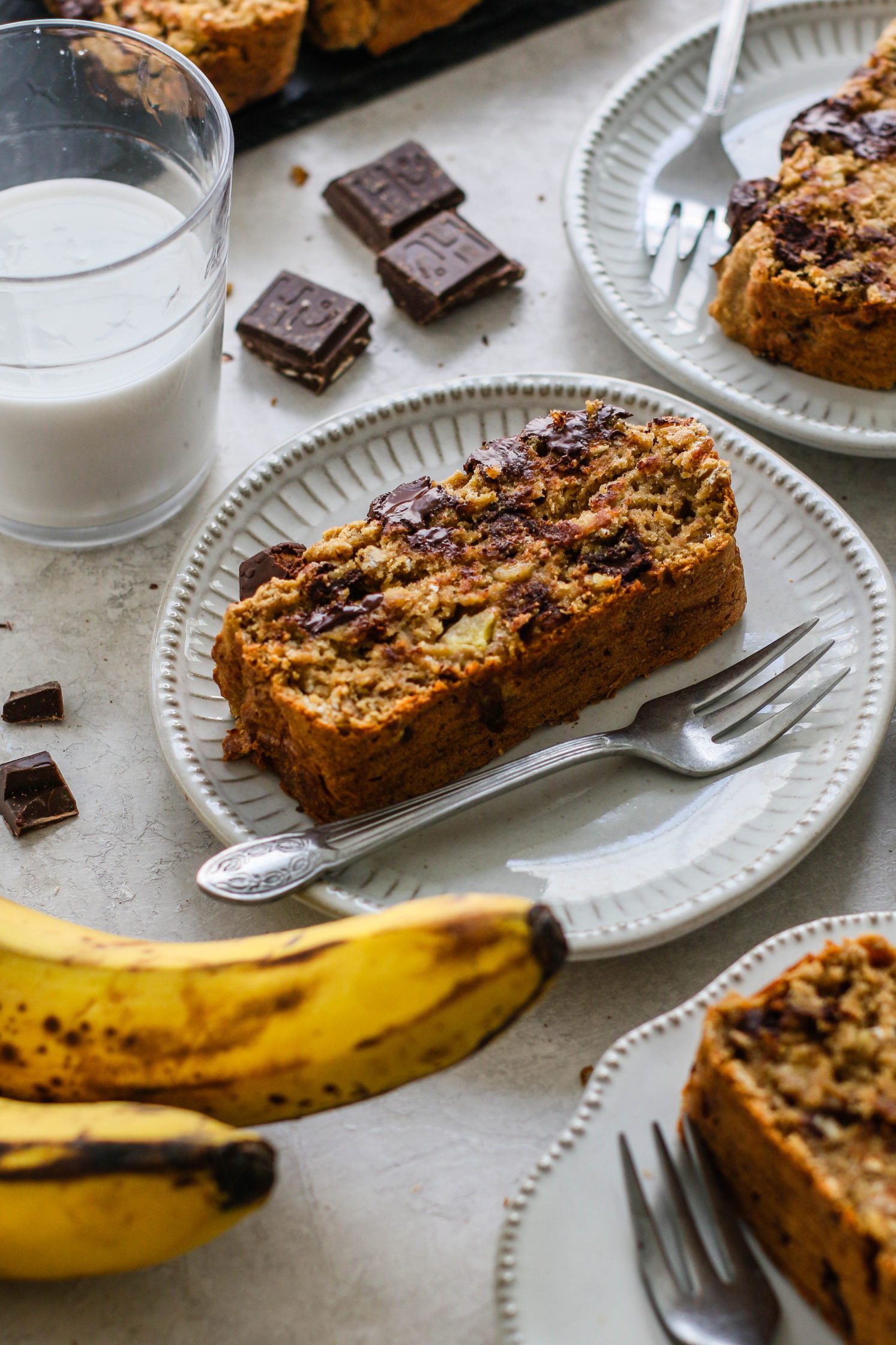 Vegan Banana Chocolate Chunk Bread served on white plates with forks with bananas and dark chocolate pieces with glass of milk