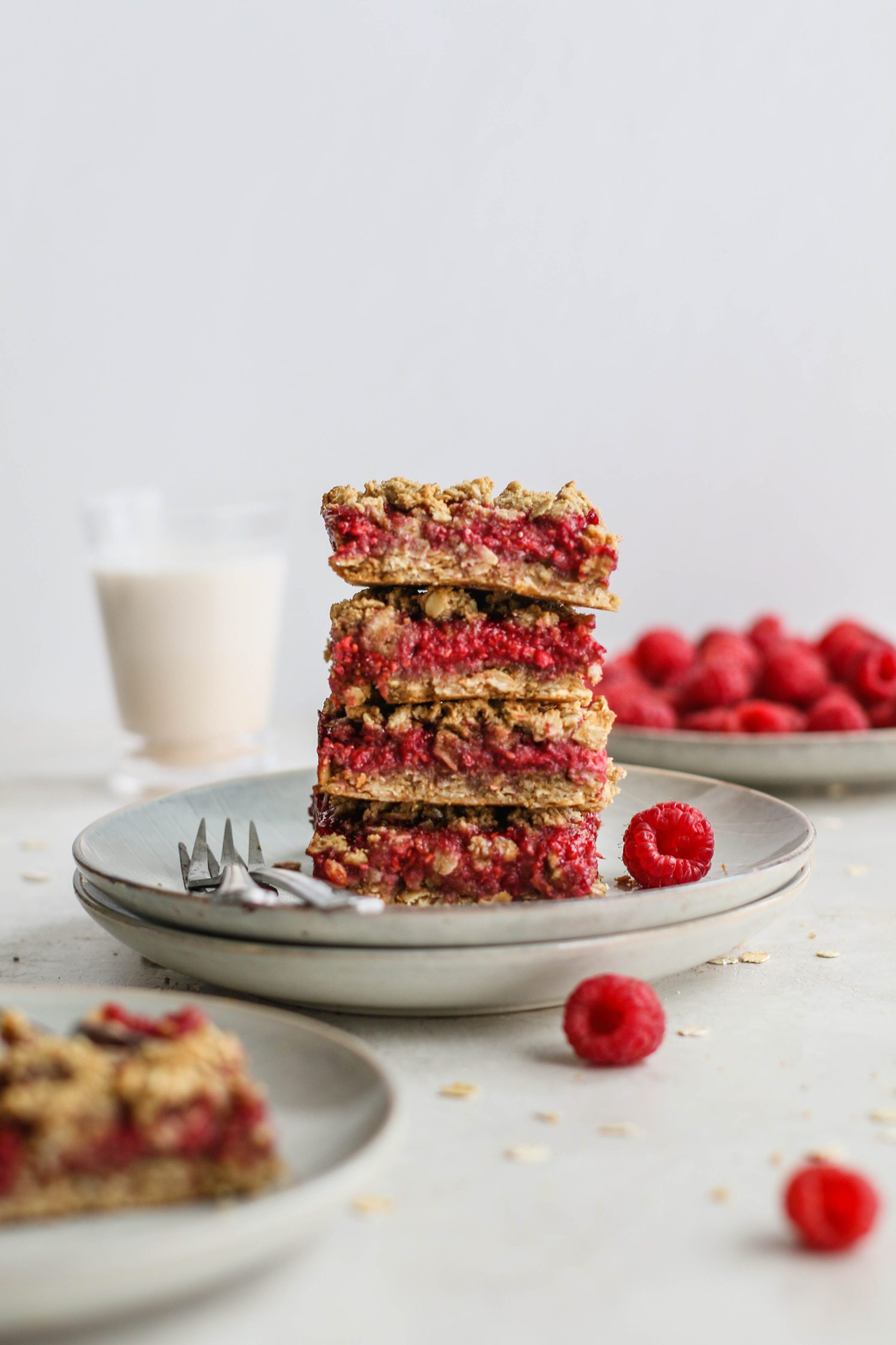 Raspberry Oat Crumble Bars stacked on two plates with fresh raspberries scattered by Flora & Vino