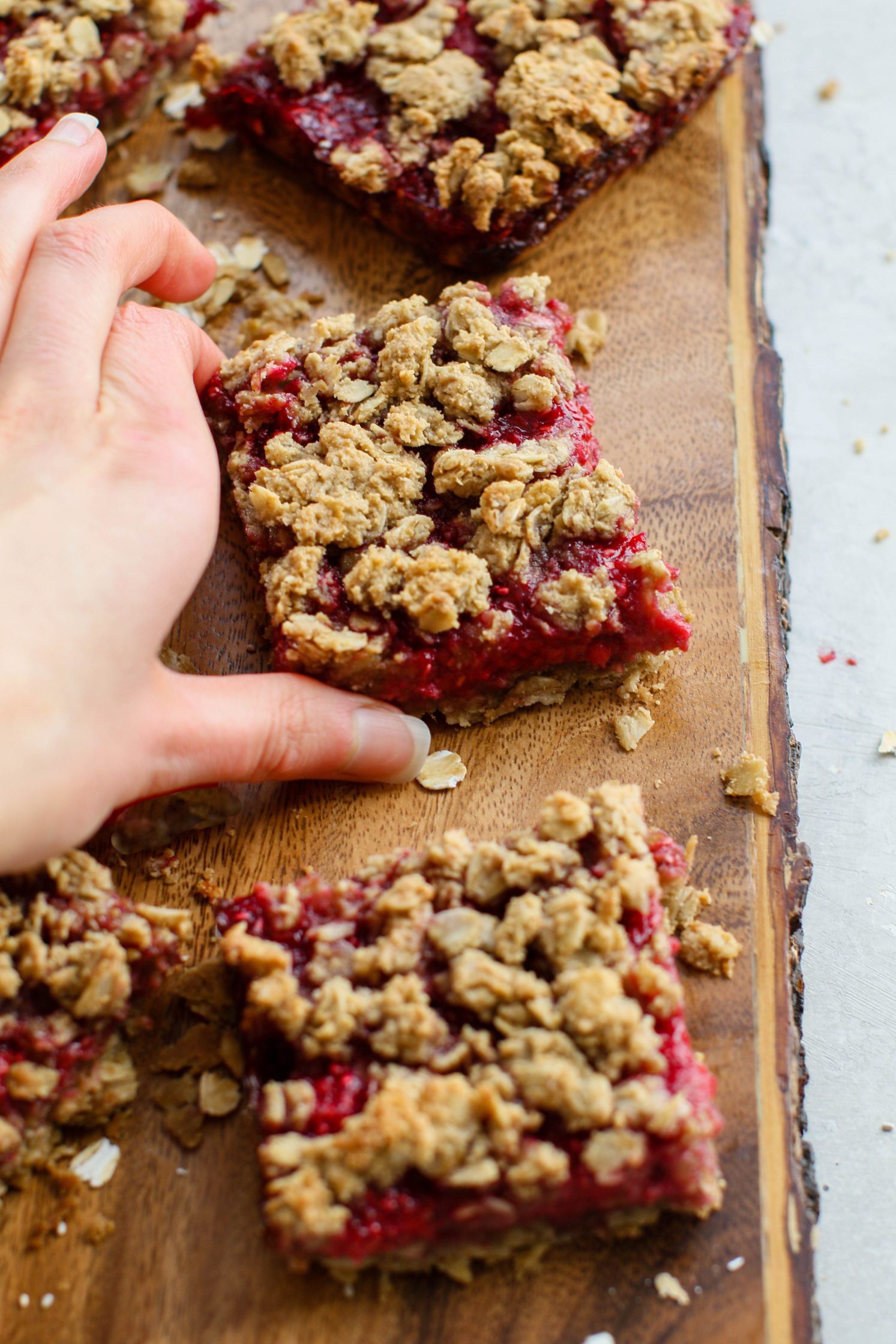 hand reaching for Raspberry Oat Crumble Bars on wooden board by Flora & Vino