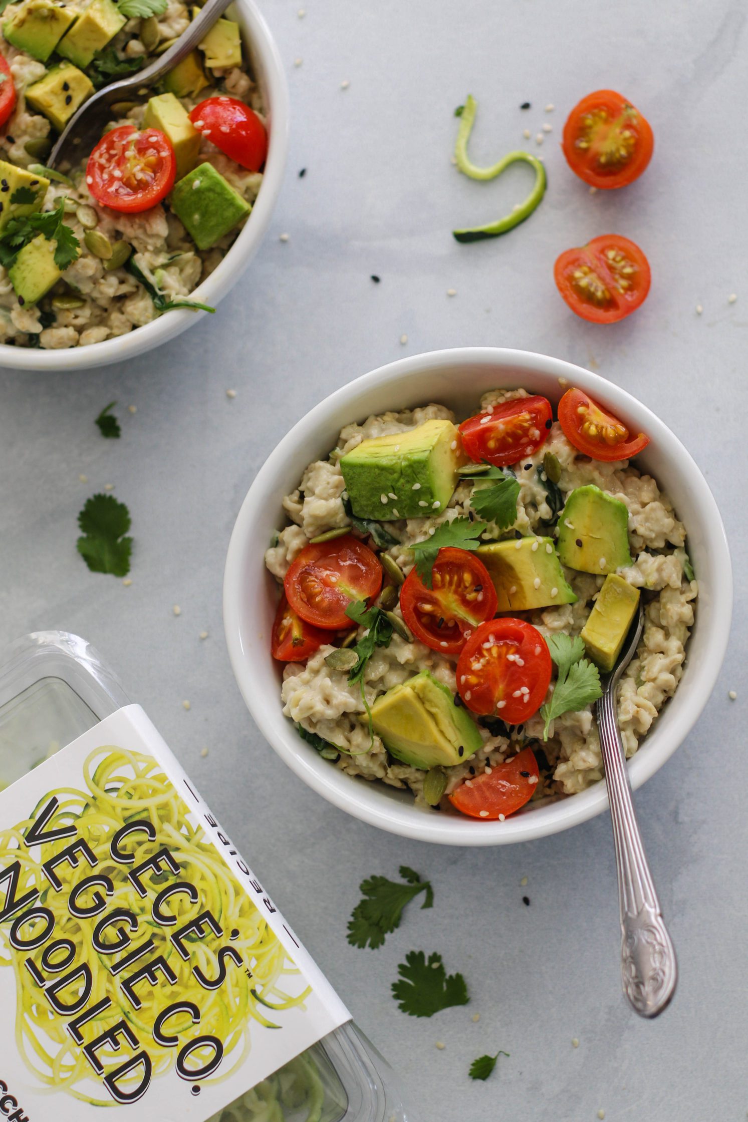 Savory Zucchini Oats (Zoats) served in bowls topped with cherry tomatoes and avocado with spoons surrounded by avocado and cherry tomatoes