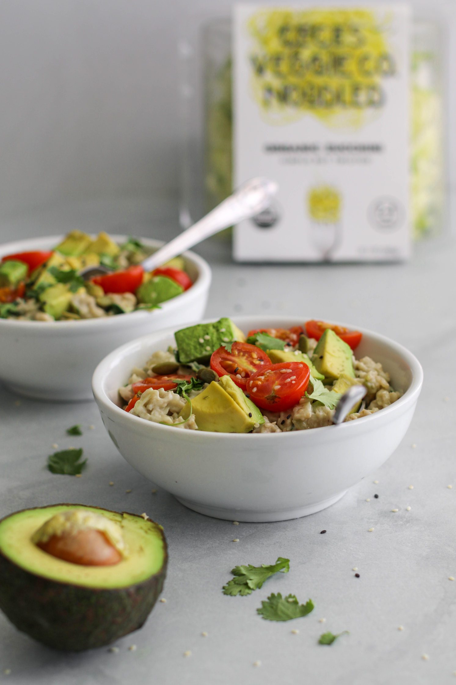 Savory Zucchini Oats (Zoats) served in bowls topped with cherry tomatoes and avocado with spoons surrounded by avocado and cherry tomatoes