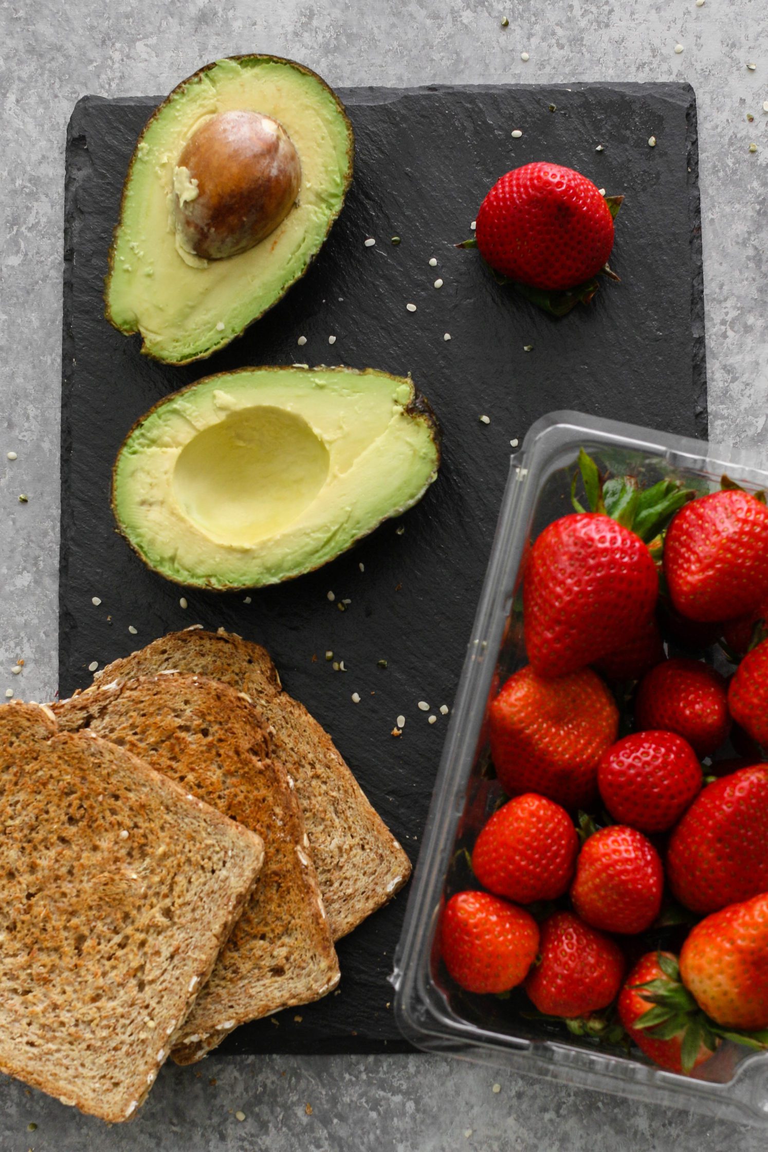 halved avocado, three slices of toasted bread, a fresh strawberries on serving board with hemp hearts sprinkled