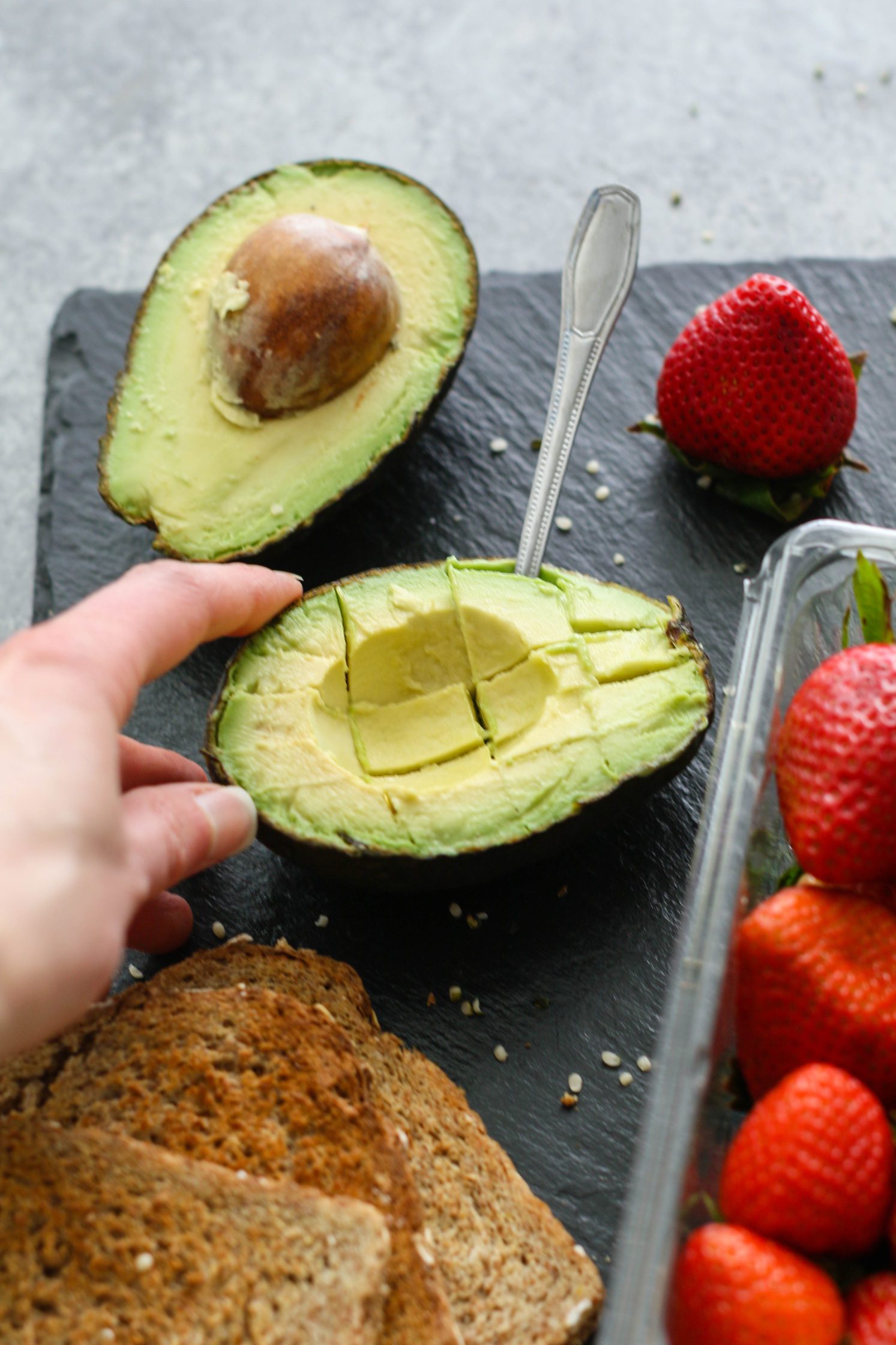 hand holding scored avocado with a spoon next to fresh strawberries and toasted breadd 