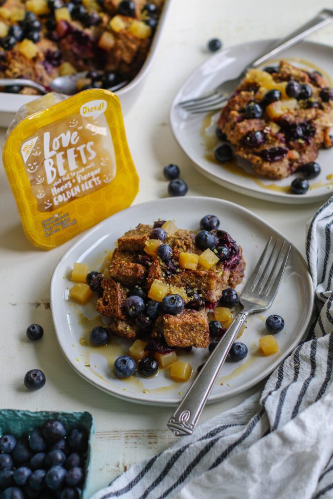 Banana Blueberry French Toast Casserole served on plate with a fork topped with fresh blueberries and golden beets with three bananas and blueberries on the side