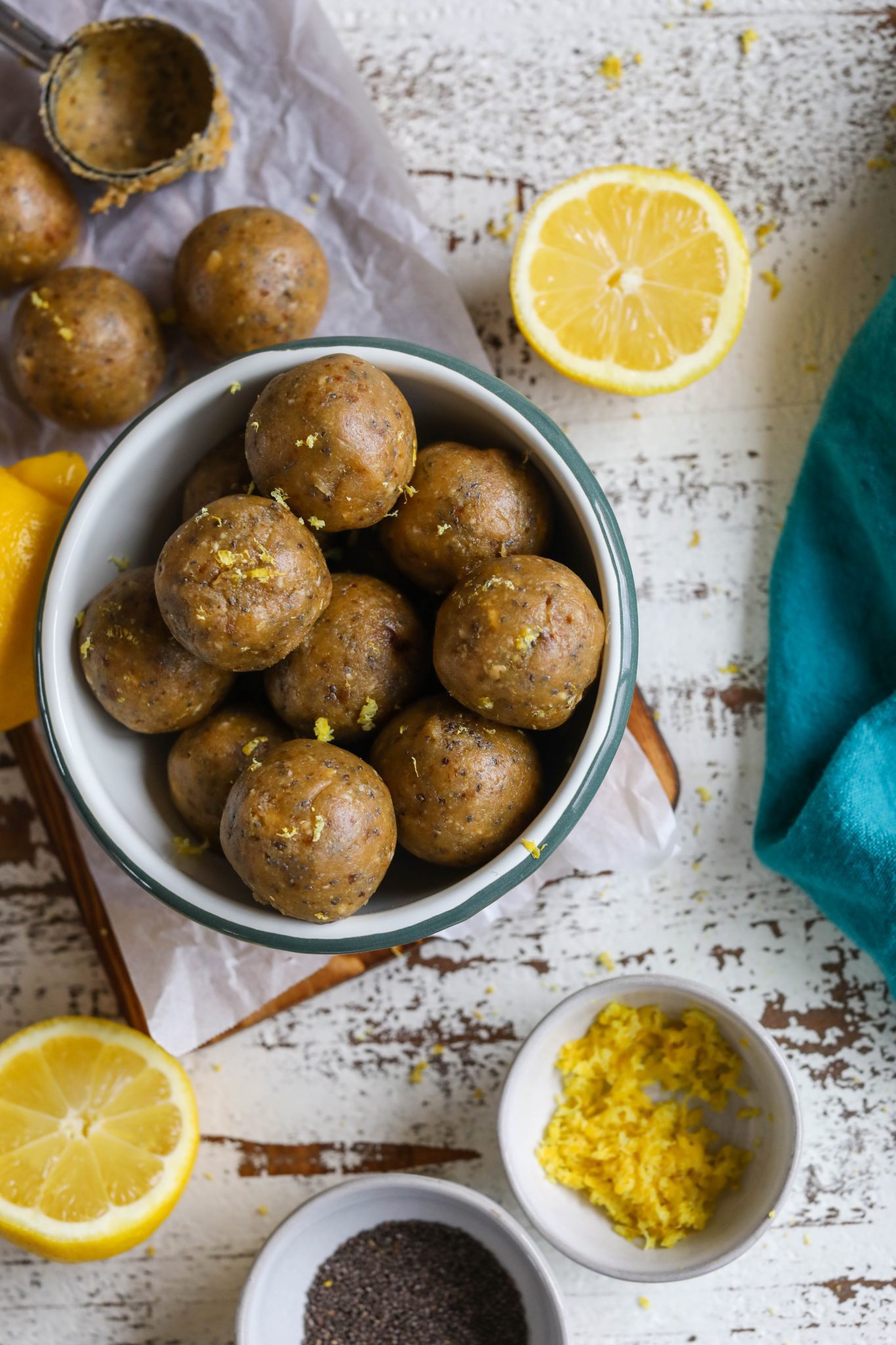 Lemon Chia Bliss Balls served in a bowl on top of parchment lined wooden board with lemons on the side, a bowl of chia seeds, a bowl of lemon zest, and a turquoise towel 