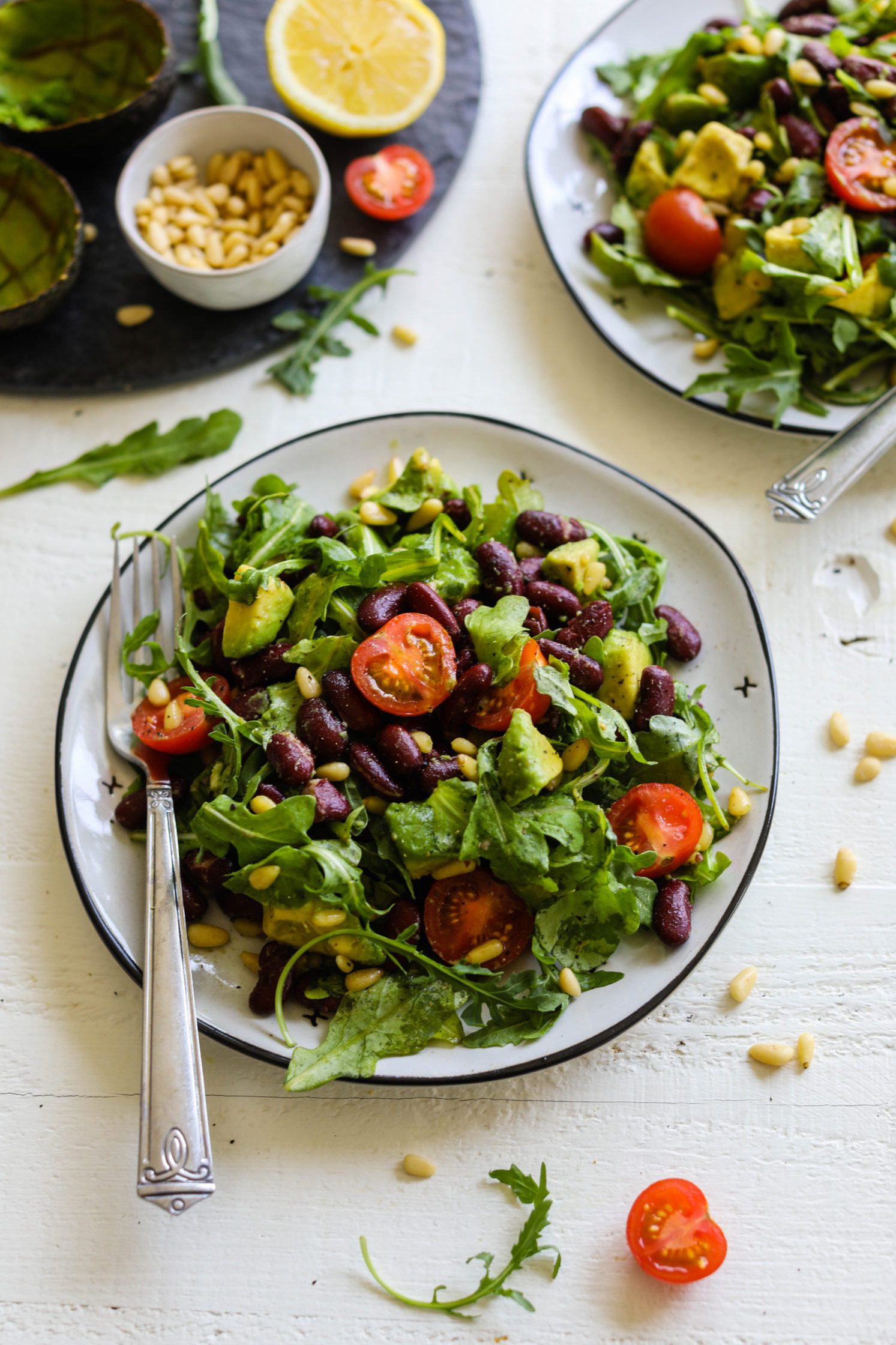 Kidney Bean Arugula Salad served on plates with forks with serving circle in corner with pine nuts, tomato, lemon, and sliced tomatoes by Flora & Vino