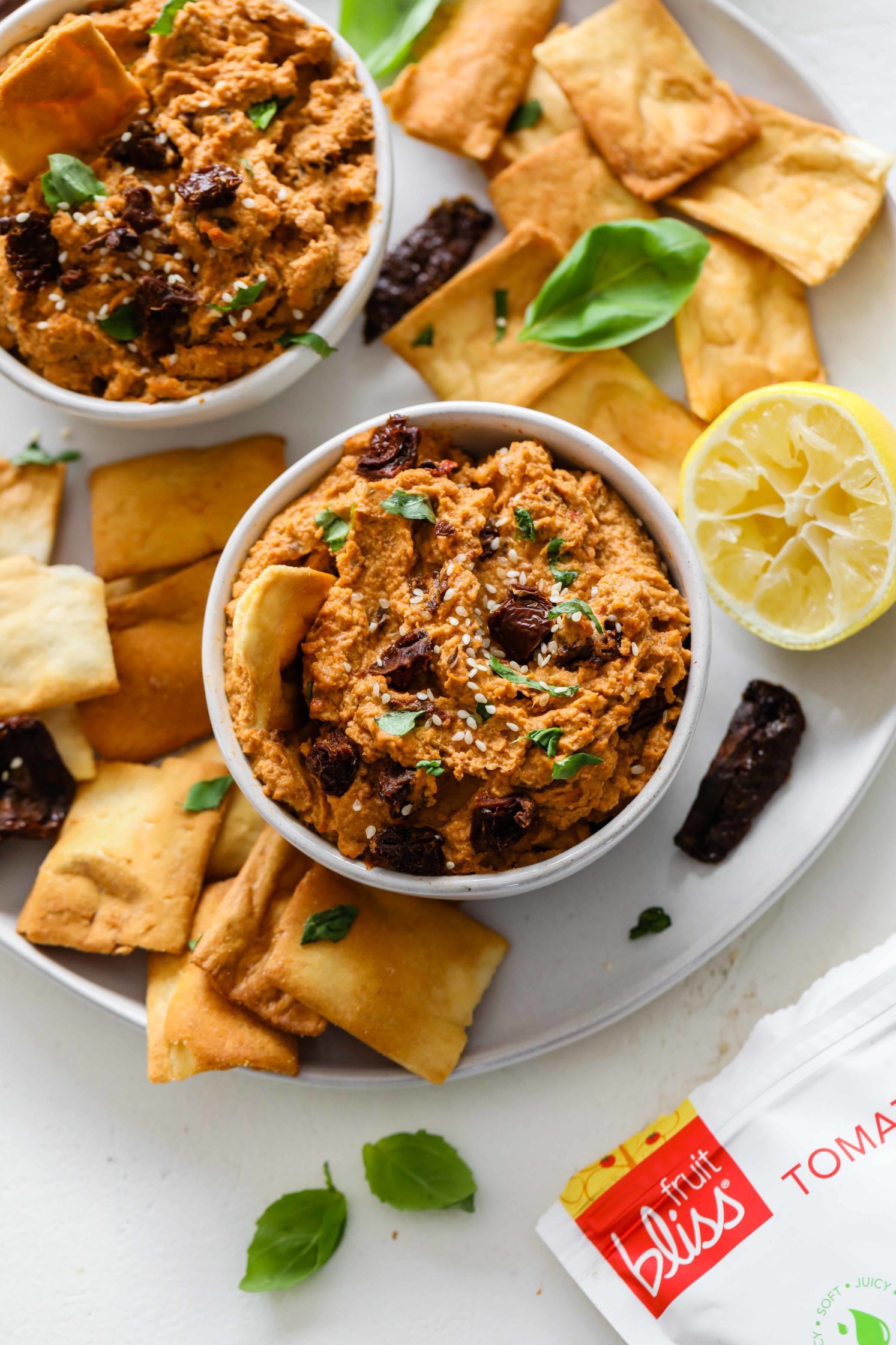 Sun-Dried Tomato & Basil Hummus served in bowls with spoons surrounded by pita chips and fresh basil and sun-dried tomato halves