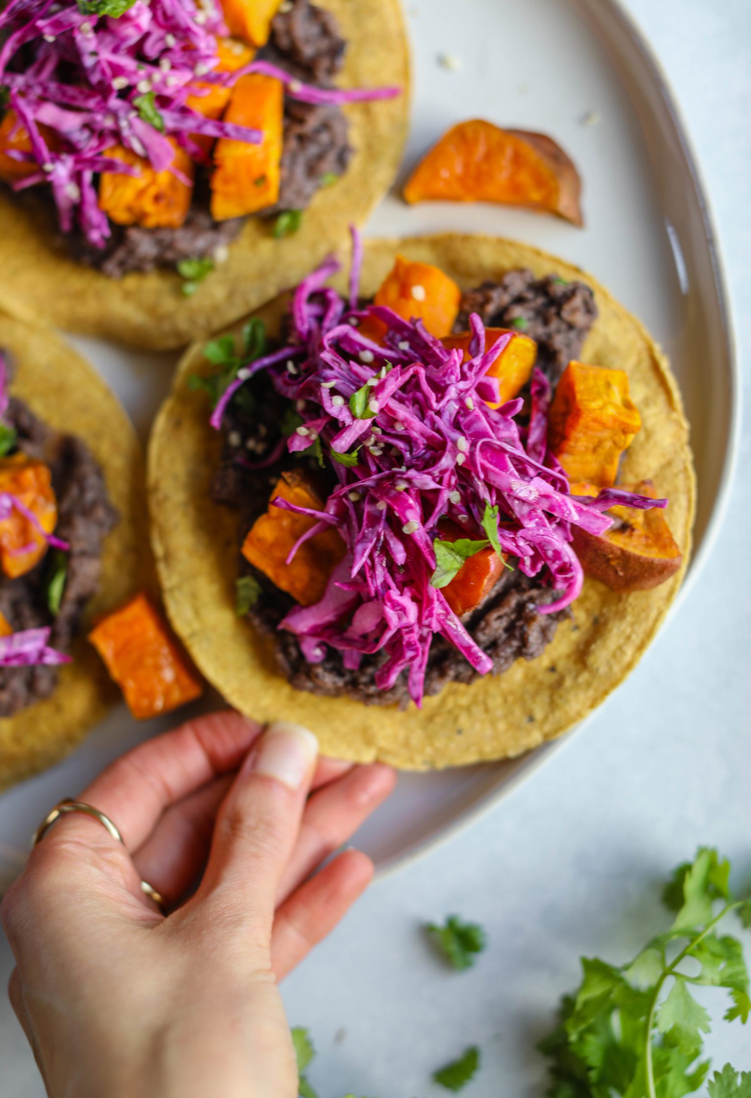 hand reaching for Sweet Potato & Black Bean Tostadas served on white plate with lime and cilantro and refried beans in pan
