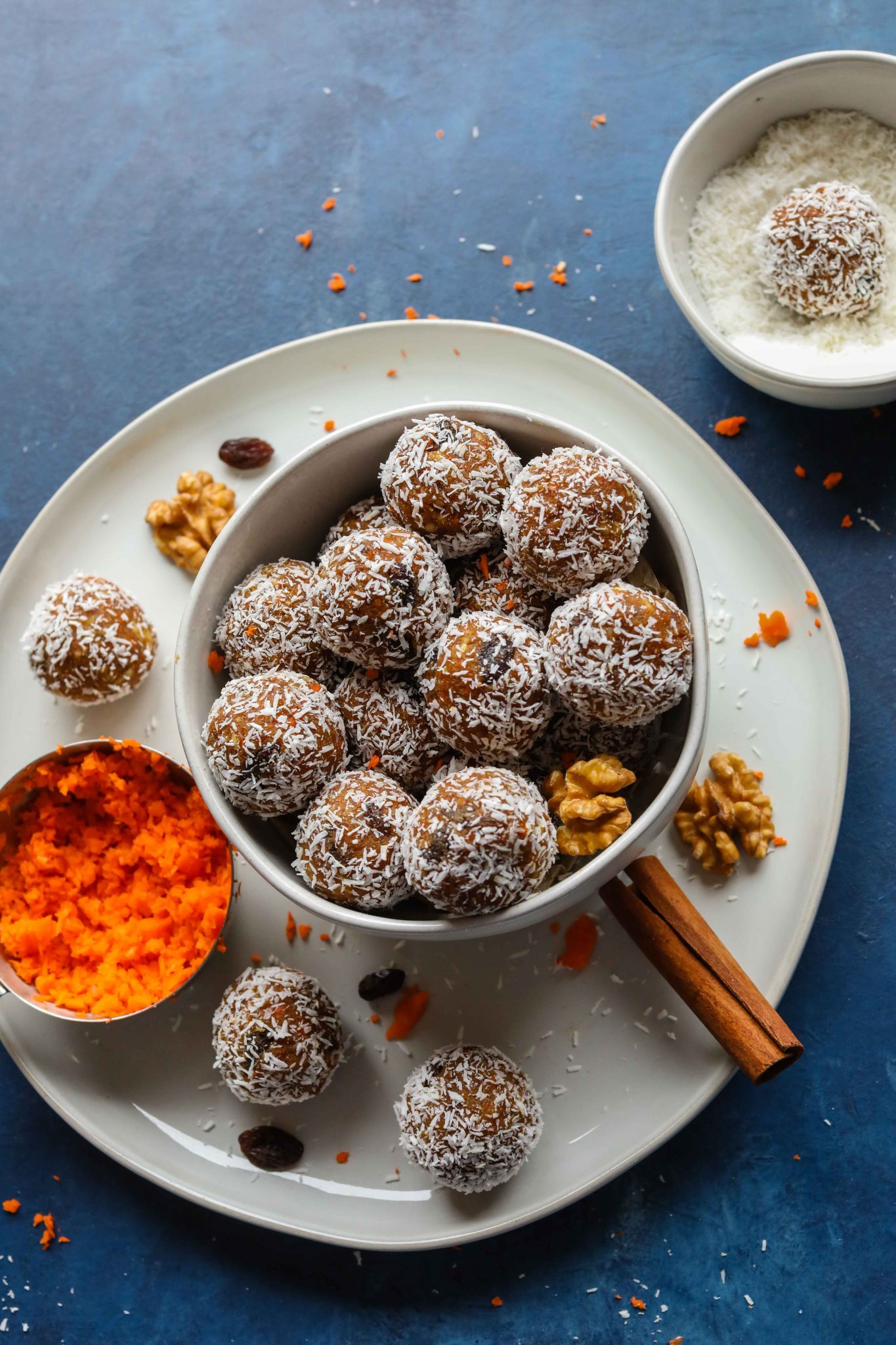 Carrot Cake Bliss Balls (Grain-Free!) served in a bowl on a plate with grated carrot, walnuts, and cinnamon sticks with a bowl of shredded coconut by Flora & Vino 