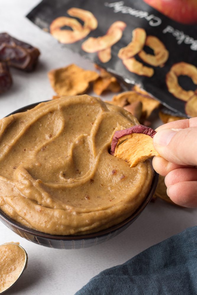 hand dipping apple chips into Tahini Date Caramel Dip by Flora & Vino