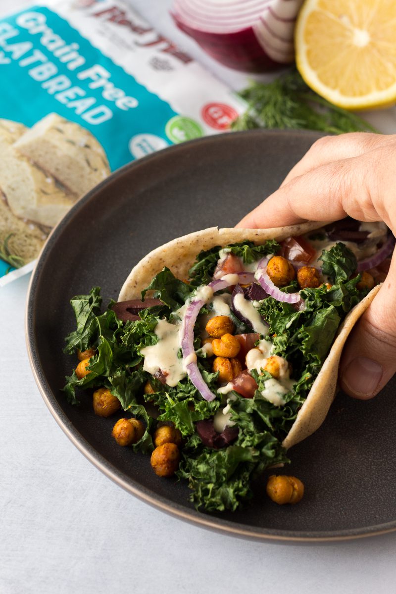 hand holding Grain-Free Flatbread Wraps served on plate with baked kale, chickpeas, red onion, and dill hummus sauce 