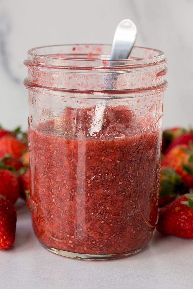 Strawberry Chia Seed Jam served in mason jar with a spoon surrounded by fresh strawberries