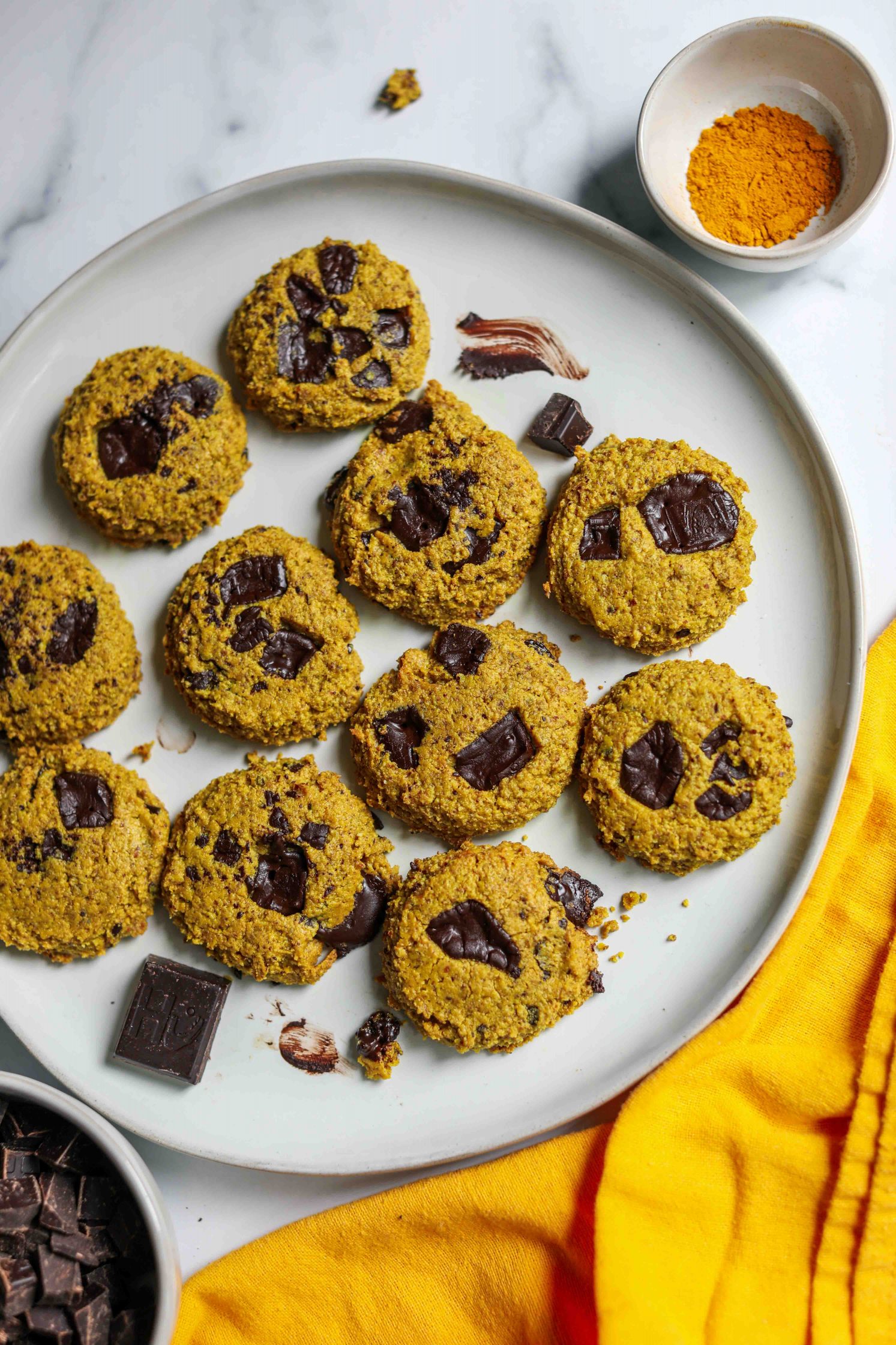 Turmeric Dark Chocolate Chunk Cookies served on a plate with a yellow towel and a side of ground turmeric and dark chocolate chunks by Flora & Vino