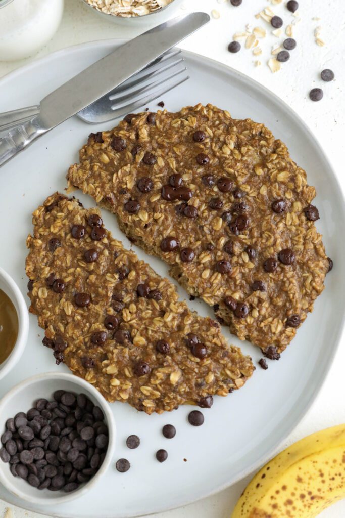 Single Banana Oatmeal Chocolate Chip Breakfast Cookie split in half on a plate with a fork and knife. There is a small bowl of chocolate chips on the plate and a banana on the side with chocoalte chips and oats scattered. 