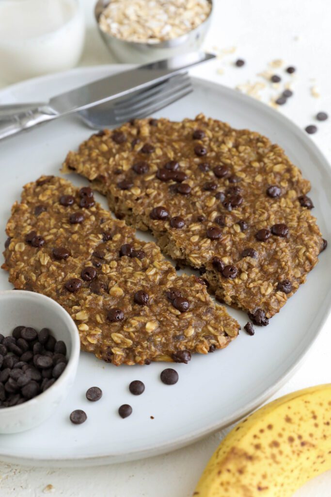 Single Banana Oatmeal Chocolate Chip Breakfast Cookie split in half on a plate with a fork and knife. There is a small bowl of chocolate chips on the plate and a banana on the side with chocoalte chips and oats scattered. 