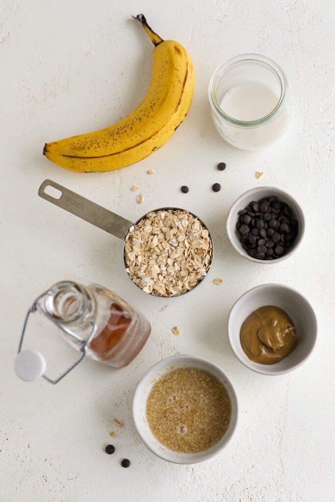white board with a glass of almond milk in a glass, mini chocolate chips in a bowl, dollop of SunButter in a bowl, flaxseed with water in a bowl, maple syrup in a container, oats in a masuring cup, and a banana.