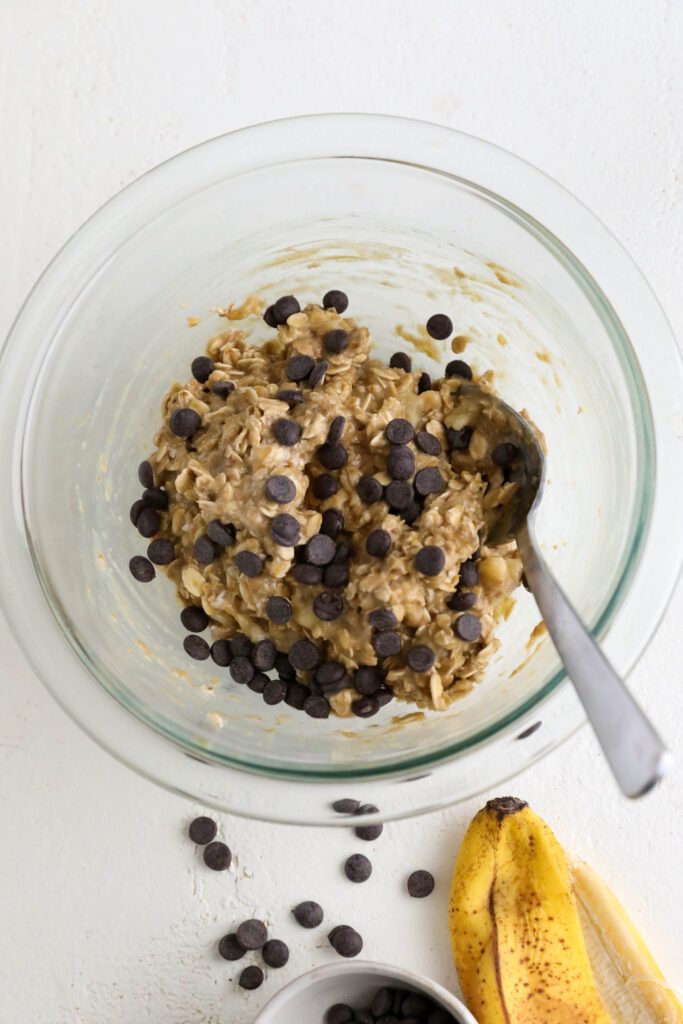 banana oatmeal cookie batter in a bowl with a spoon with dark chocolate chips scattered on top. There is a banana peel and scattered dark chocolate chips on the bottom right.