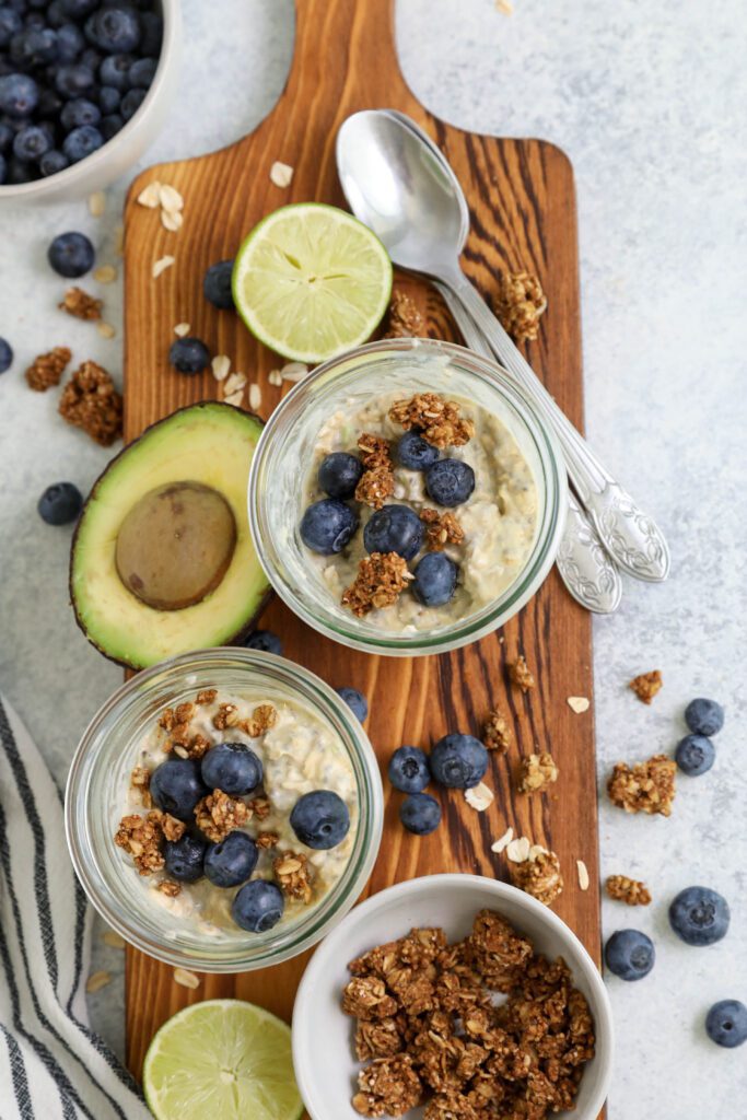 Key Lime Pie Overnight Oats served in mason jars on a wooden board topped with blueberries and granola with two spoons by Flora & Vino