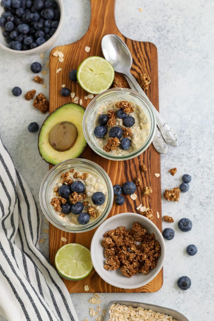 Key Lime Pie Overnight Oats served in mason jars on a wooden board topped with blueberries and granola with two spoons by Flora & Vino