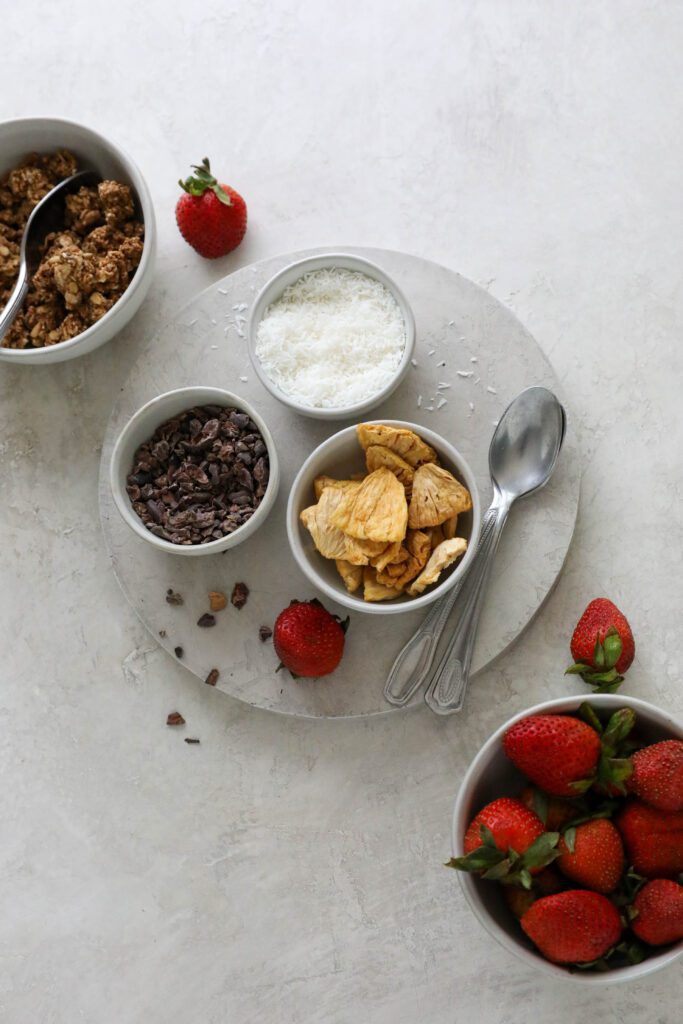 granola, cacao nibs, shredded coconut, and dried pineapple on circle with two spoons and fresh strawberries