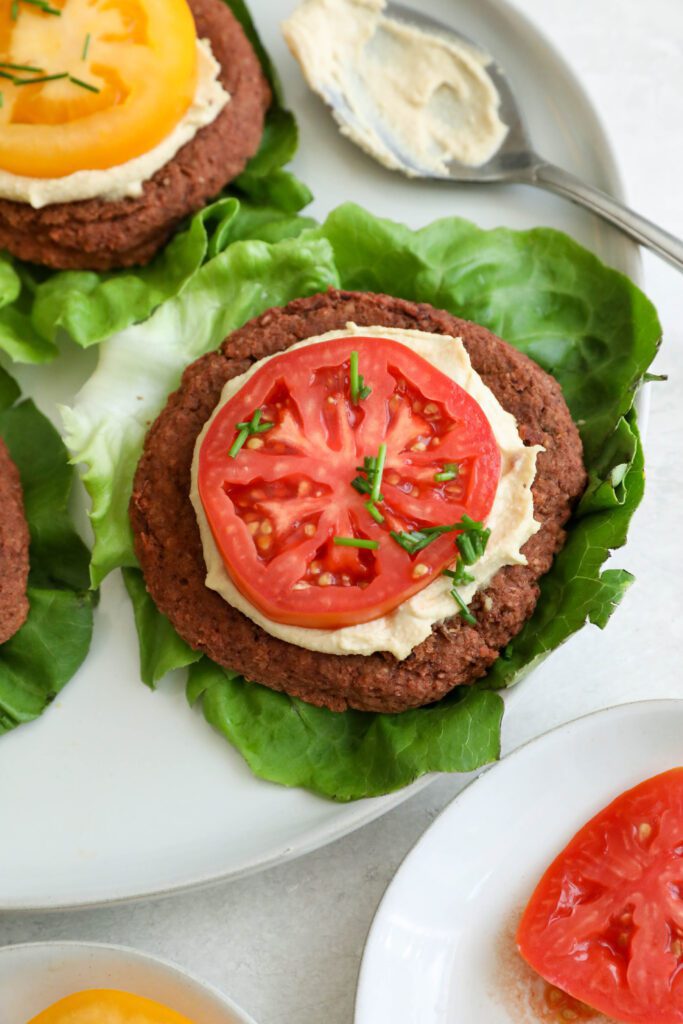 Smoky White Bean & Beet Burgers served on lettuce leaves on white plate topped with hummus and tomato