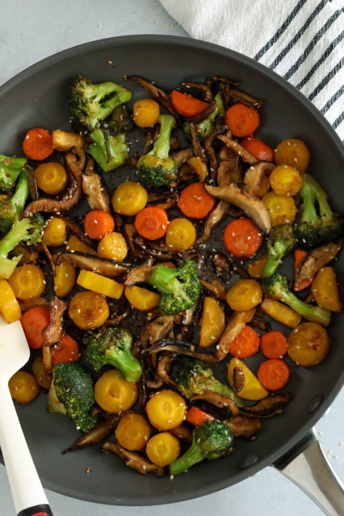 Sesame Vegetables in pan topped with sesame seeds and spatula with a blue and white striped towel in corner by Flora & Vino
