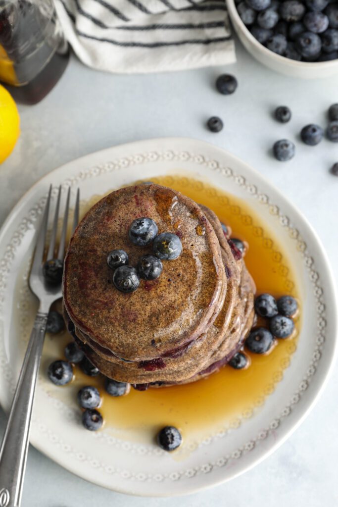 Blueberry Buckwheat Pancakes stacked on plate served overhead with blueberries and a fork by Flora & Vino