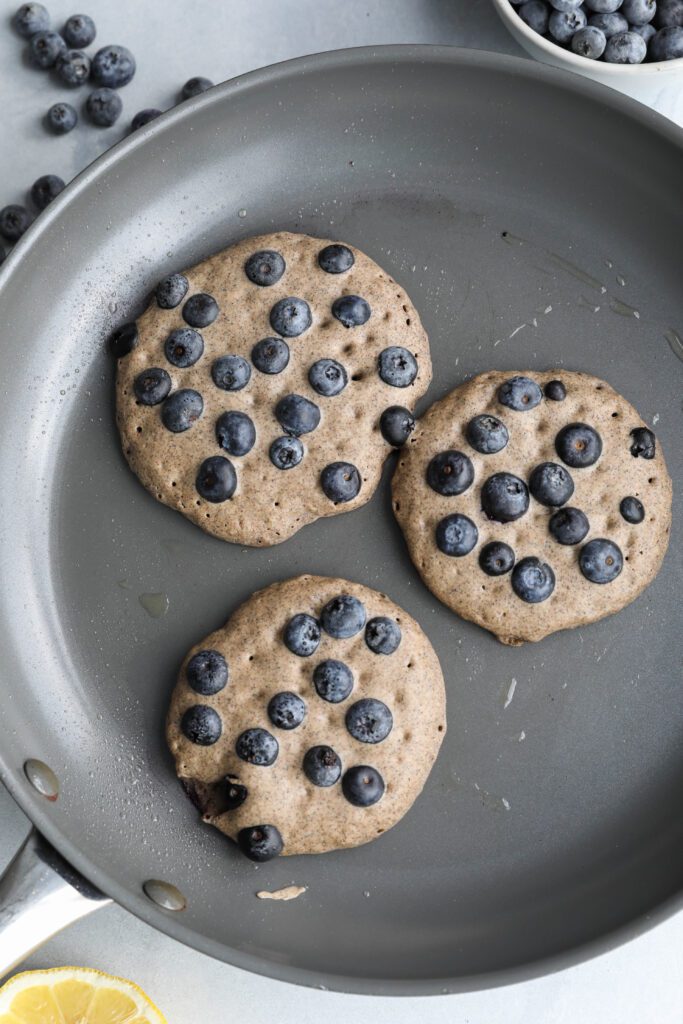 Blueberry Buckwheat Pancakes formed in pan with blueberries on top with blueberries and lemon on the side by Flora & Vino