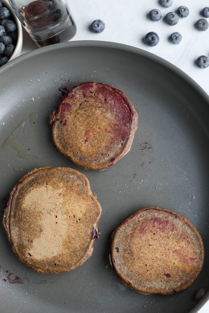 Blueberry Buckwheat Pancakes flipped in pan with blueberries on the side by Flora & Vino
