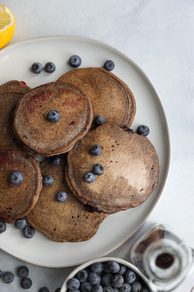 Blueberry Buckwheat Pancakes on plate with blueberries sprinkled on top with lemon on the side by Flora & Vino