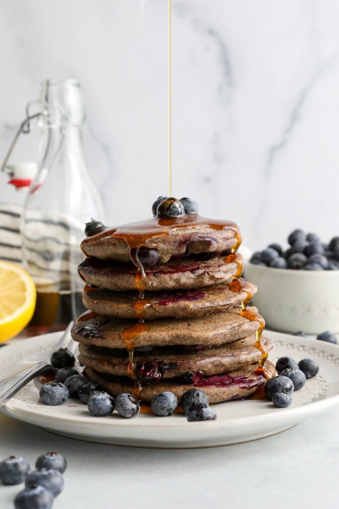 Blueberry Buckwheat Pancakes stacked on a plate with blueberries and maple syrup drizzled on top by Flora & Vino