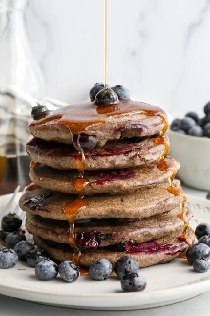 Blueberry Buckwheat Pancakes stacked on a plate with blueberries and maple syrup drizzled on top by Flora & Vino