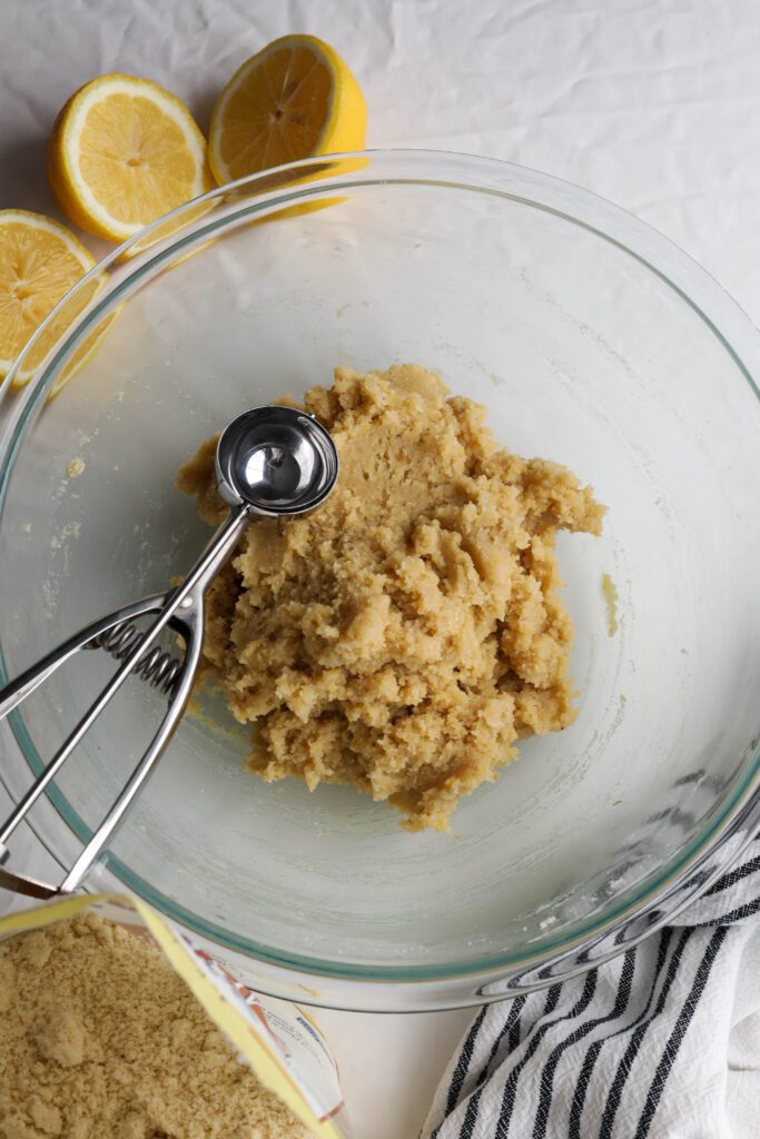 Lemon Almond Flour cookie batter in glass bowl with halved lemons and almond flour on the side with cookie dough scoop