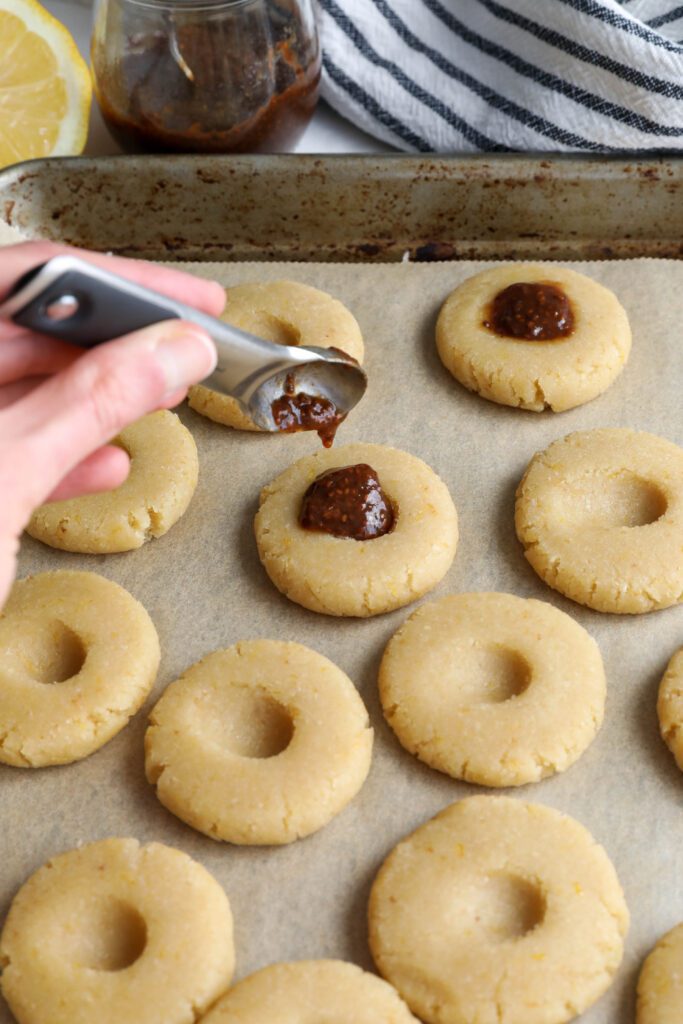 hand adding jam to center on lemon almond flour thumbprint cookies on parchment lined baking sheet