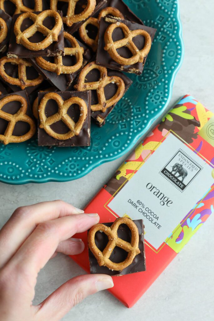 Orange Chocolate Pretzel Bark served on blue plate with hand holding one piece on top of chocolate bar by Flora & Vino