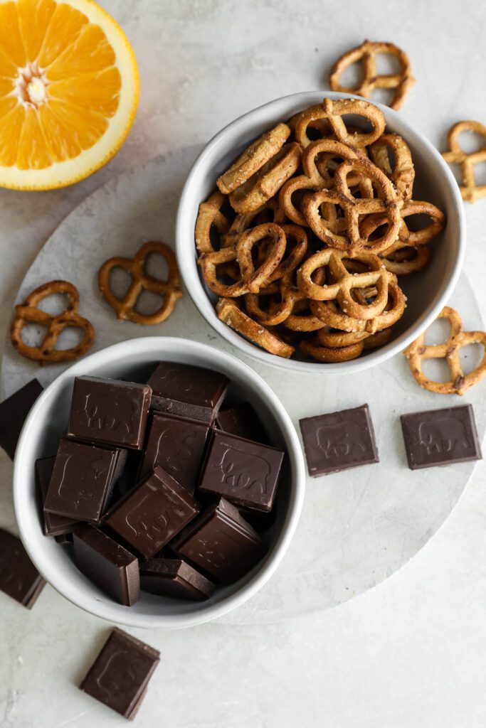 pretzels in a bowl, chocolate squares in a bowl, and orange halve on white circle by Flora & Vino