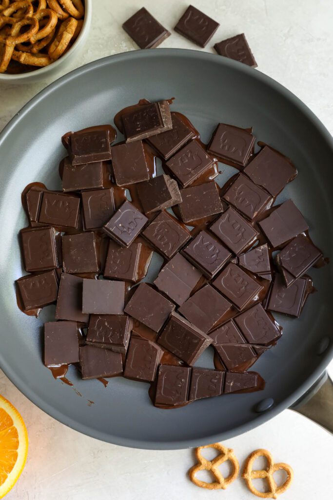 chocolate squares melting in pan with pretzels and orange and shopped chocolate and pretzels around it by Flora & Vino