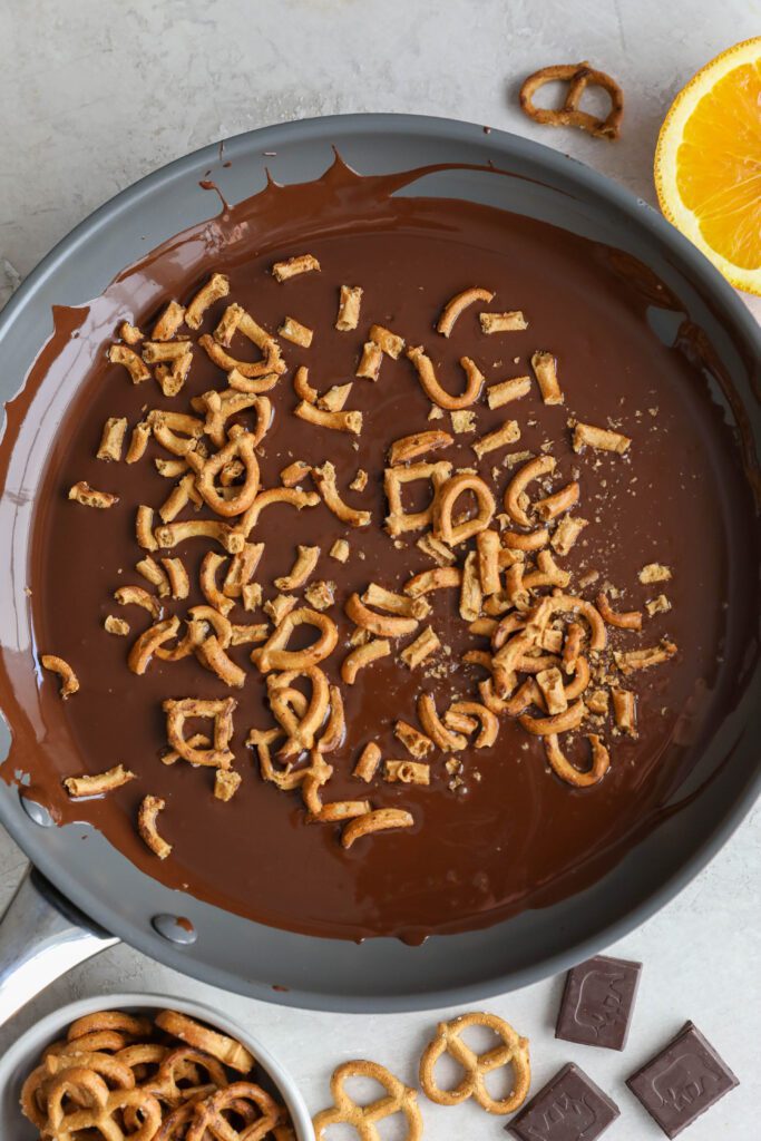 chopped pretzels in melted chocolate in saucepan with pretzels and chocolate squares and orange on the side by Flora & Vino