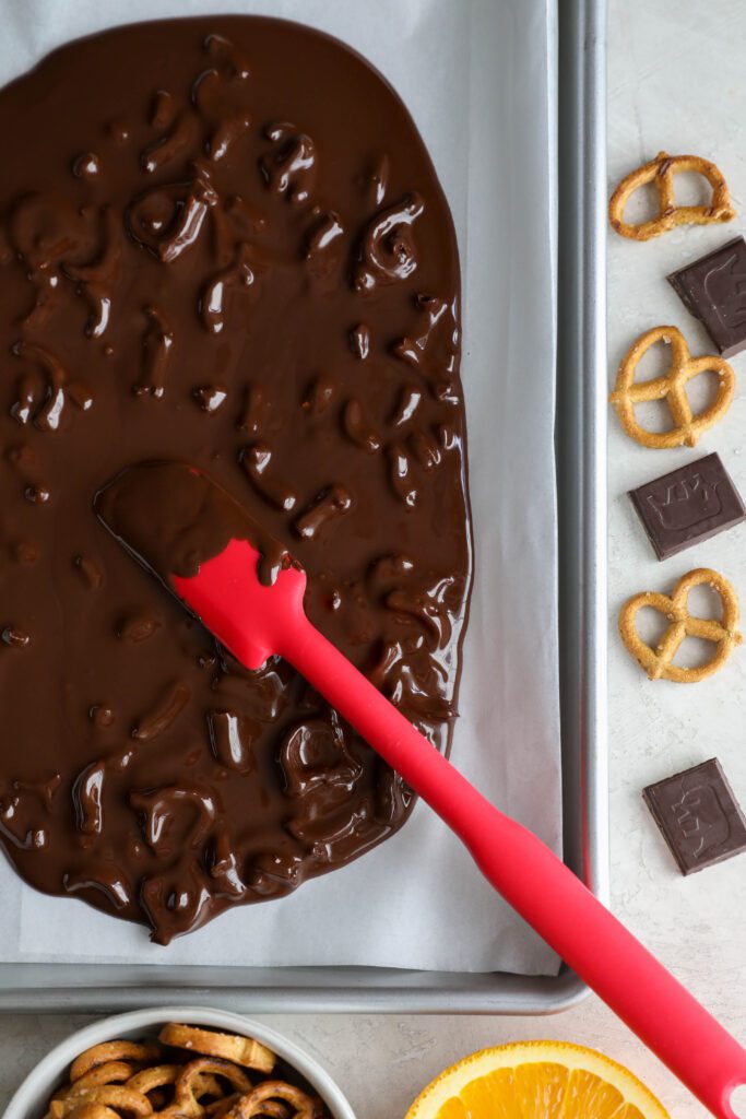 red spatula spreading melted chocolate with pretzels on parchment lined baking sheet with pretzels and chopped chocolate and orange on the side by Flora & Vino