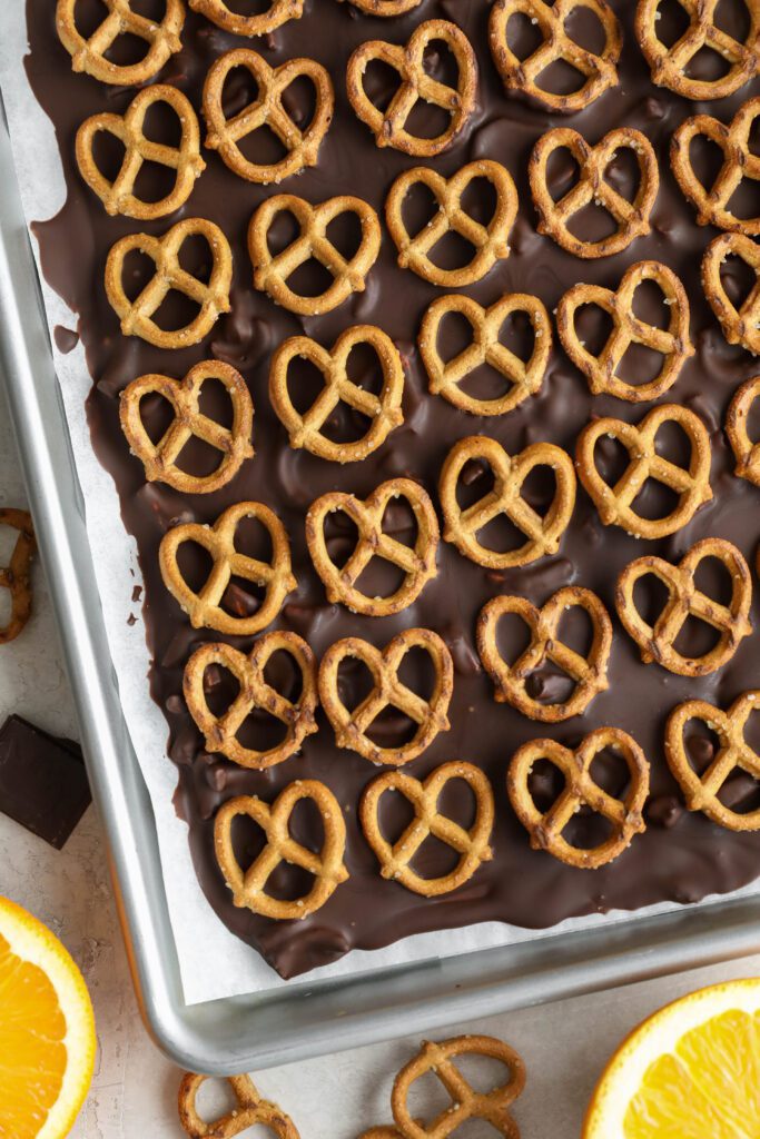 Orange Chocolate Pretzel Bark frozen on parchment lined baking sheet with pretzels and oranges and chocolate on the side by Flora & Vino