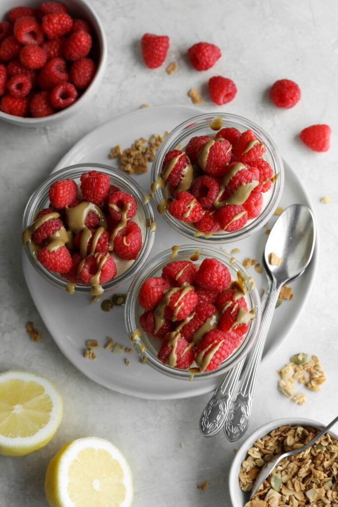 SunButter Cheesecake Cups topped with fresh raspberries and SunButter on plate with spoons with lemon and fresh raspberries and granola on the side