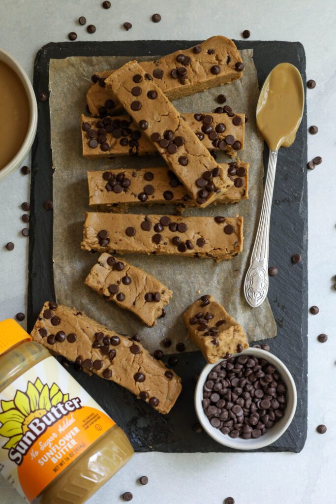SunButter Chocolate Chip Protein Bars on parchment lined board with a SunButter spoon and a bowl of chocolate chips by Flora & Vino